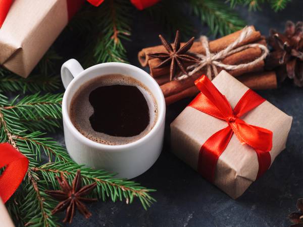 En kopp med julekaffe omgitt av julepynt og gaver, med granbar og krydderstjerne rundt.