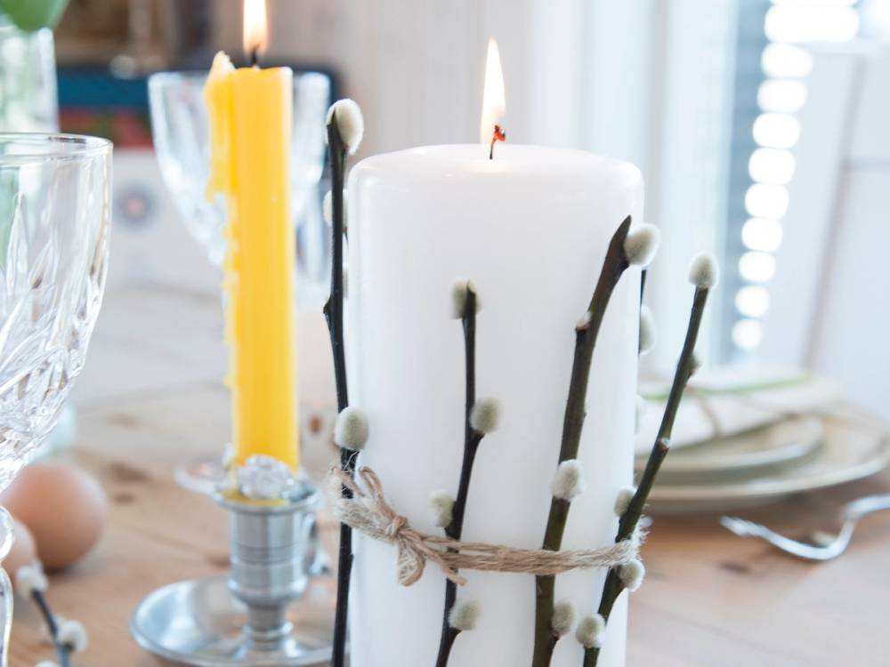 Påskebord med pyntet hvit stor lysestake og gule lys, omgitt av andre lys og borddekking.