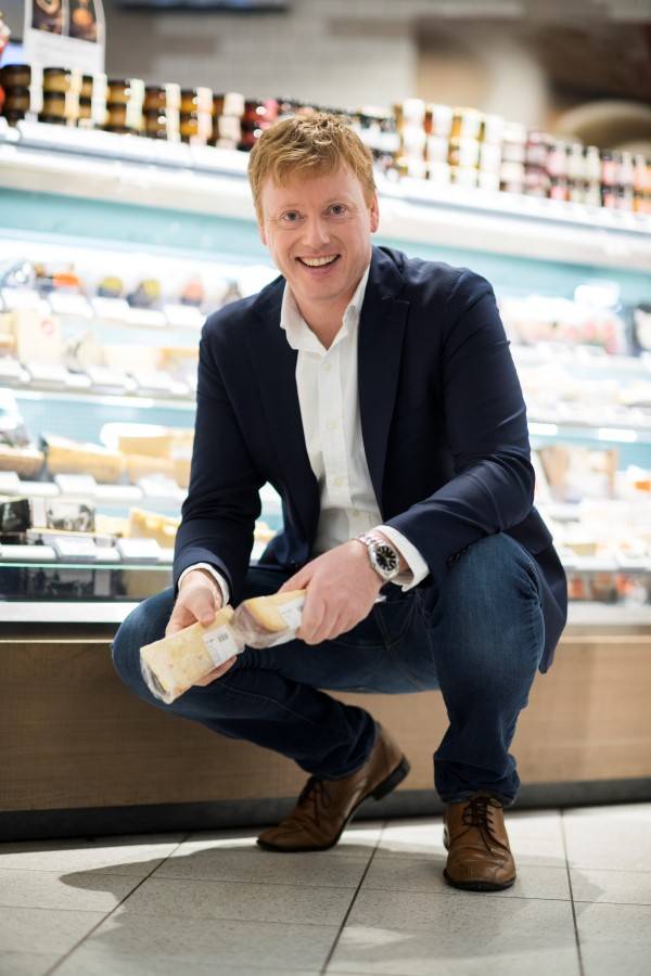 En mann i dress og hvit skjorte som holder en ostebit i en butikk med kjølehyller fylt med meieriprodukter.
