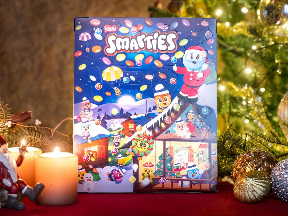 En julekalender med Småties-logo og julemotiver står på et bord med lys og julepynt rundt, foran et juletre.