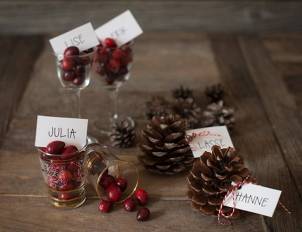 Julekort og navnelapper med julepynt og kongler på et treverkebord, med navn som Lise, Julia og Hanne på kortene.