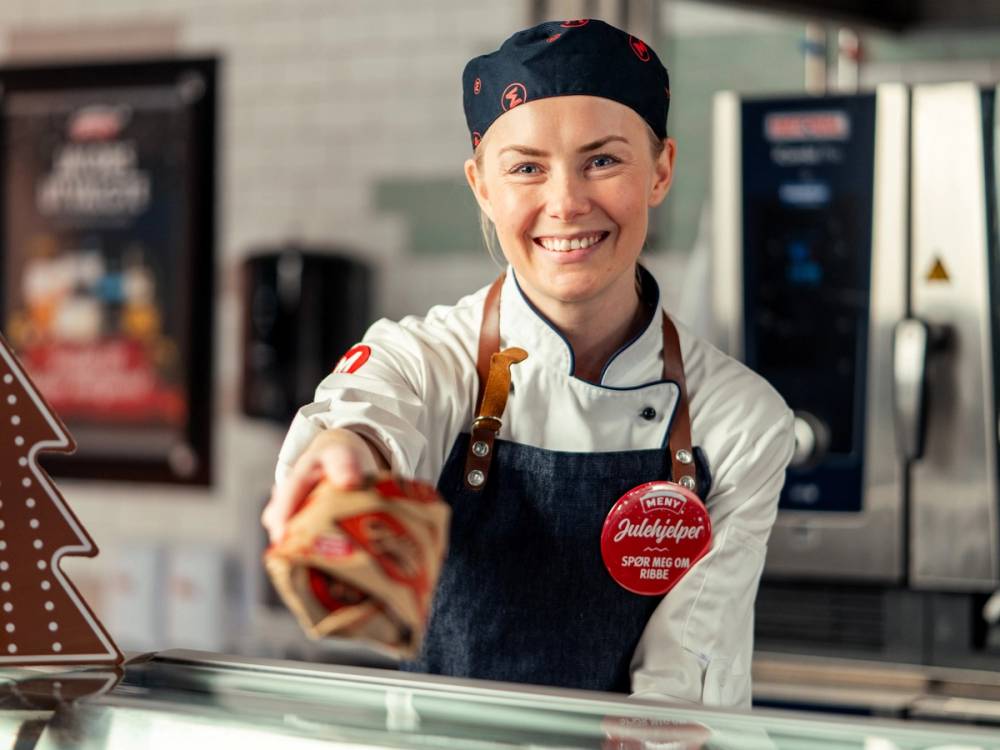 En person i arbeidstøy som serverer mat i en butikk eller kafé, med et forkle og en knapp med tekst.