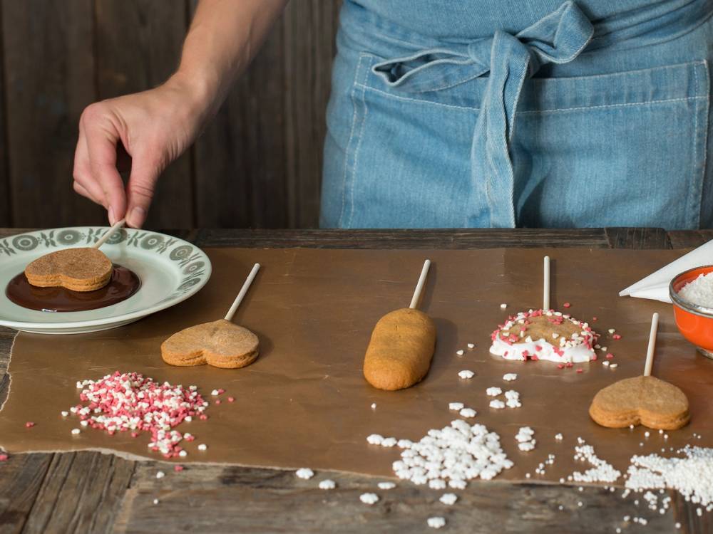 Person som lager kjeks med ulike dekorasjoner på et kjøkkenbord, med en hånd som holder en kjeks på en tallerken.