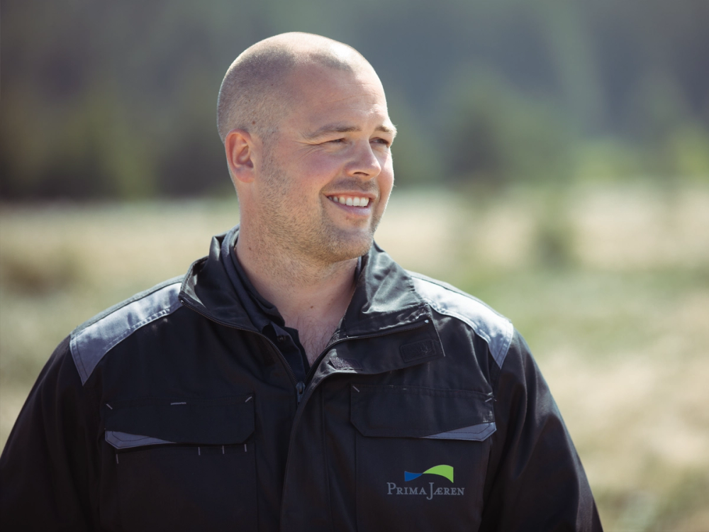 En person med kort hår og mørk jakke står utendørs med en uskarp naturbakgrunn.