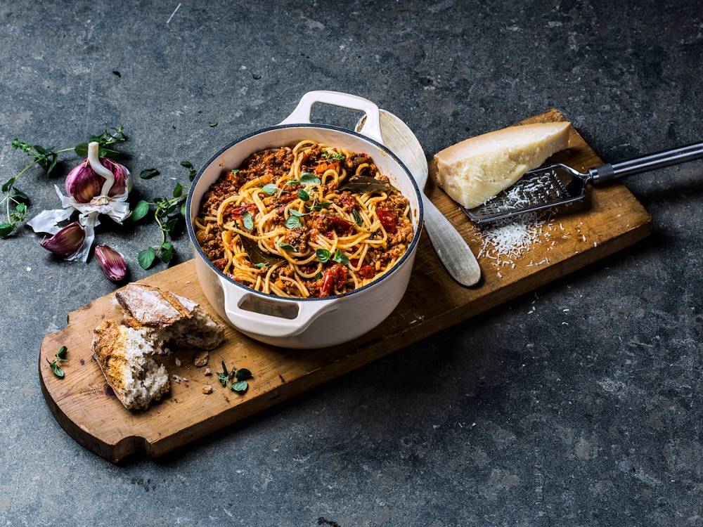 En bol med pasta bolognese på et trebrett, omgitt av hvitløk, urter og et stykke brød, på en mørk bakgrunn.