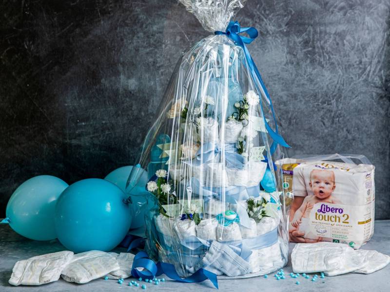 En bleiekake dekorert med hvite blomster og blå bånd, omgitt av blå ballonger og en pakke med bleier, sannsynligvis til en babyfeiring.