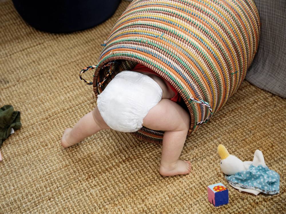 Et barn som har hodet og armer inne i en oppbevaringspose eller sekk, med kroppen utenfor, på et teppe.