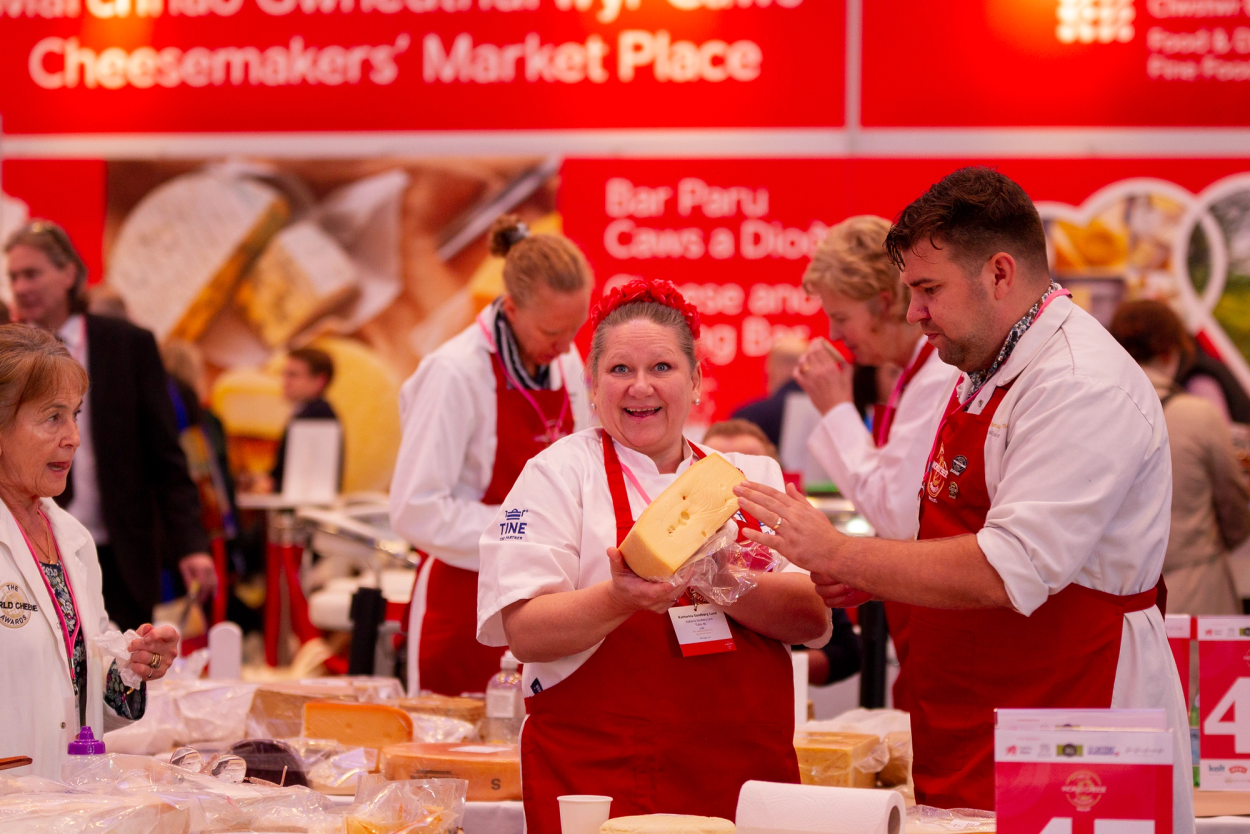 Tre personer i hvite klær og røde forkle som ser på en ost på en matmesse eller utstilling, med en bakgrunn av matrelaterte plakater og skilt.