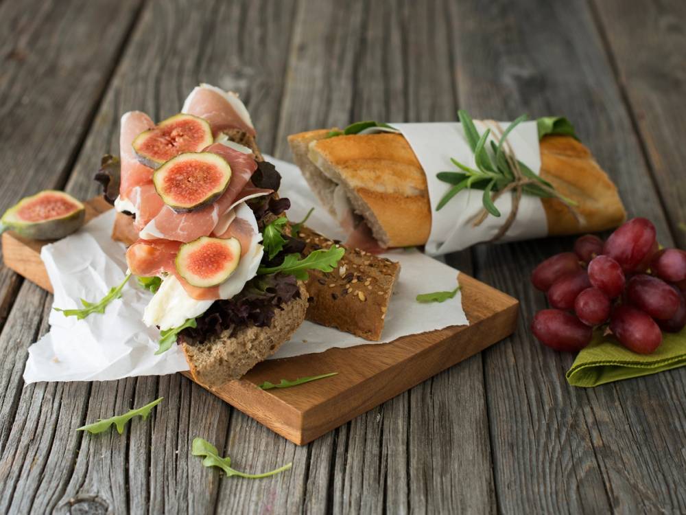 En baguette fylt med spekeskinke og brieost, servert på et trebrett med druer ved siden av.