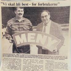 To personer står sammen og holder opp et stort skilt med ordet 'MENY' på, foran en avisutklipp som viser en artikkel om meny og priser.