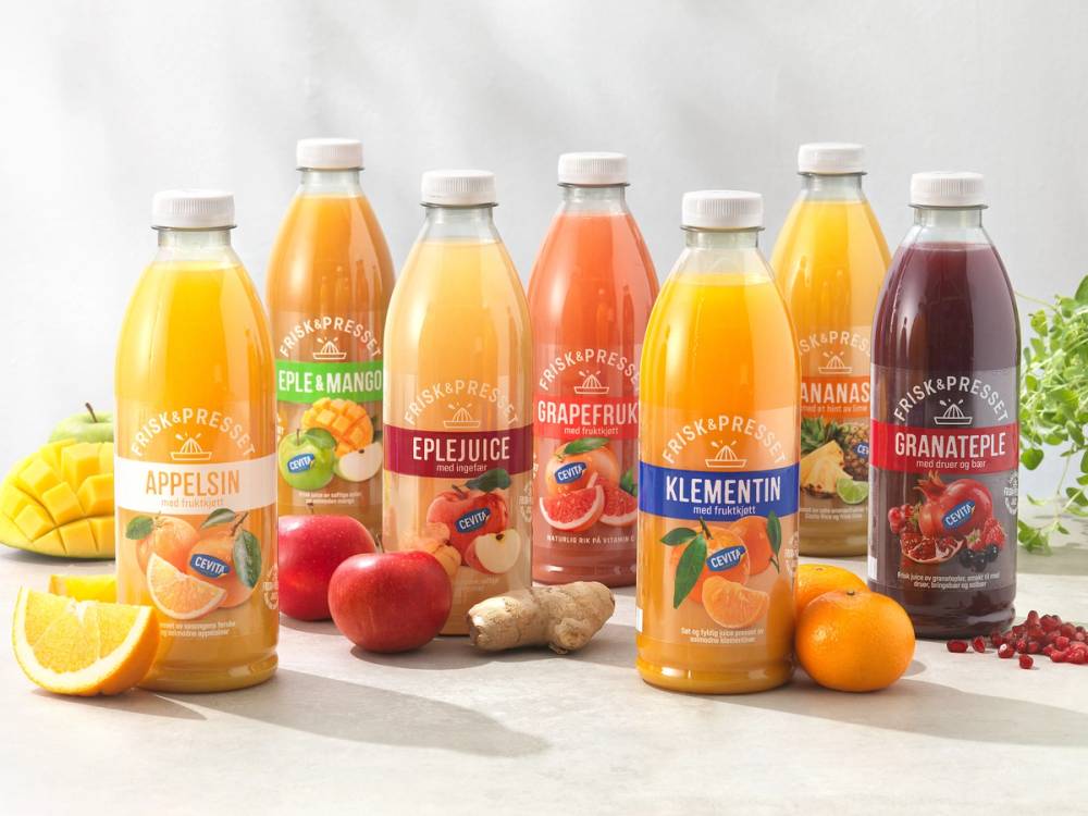 Syv flasker med ulike typer fruktjuice, inkludert appelsin, eple, grapefrukt, klementin, granateple og en annen type, plassert på et bord med frukt og urter rundt.