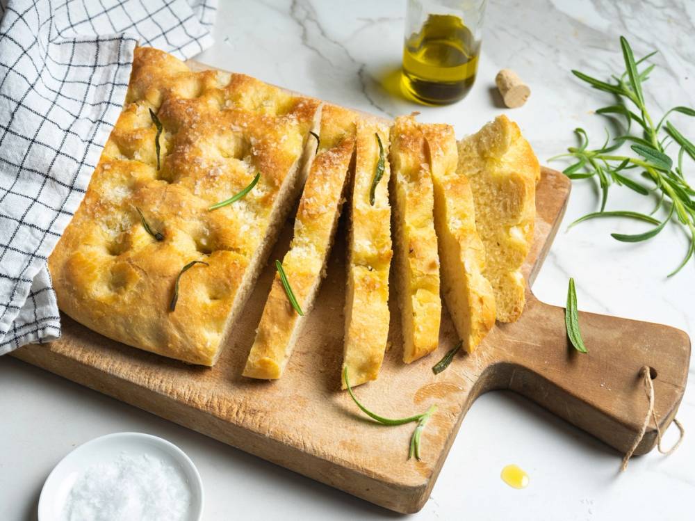 Focaccia med sprø skorpe og urter, skåret i biter på et skjærebrett med olivenolje og urter i bakgrunnen.