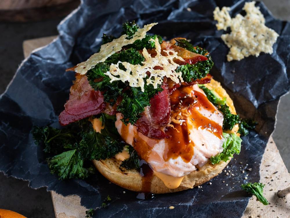 Burger med parmesanchips på toppen, ser ut til å inneholde egg, kjøtt, grønnsaker og saus, på en svart bakgrunn.