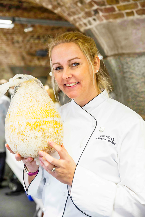 Kvinne i kokkeforkle med navnemerke som sier Siri-Helen Hansen-Barry holder en stor ost i en bakke, med en steinbro i bakgrunnen.