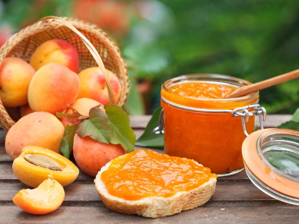 Fersken og aprikoser i en kurv med et glass med aprikossyltetøy og en skive brød med syltetøy på en utendørs trebord.