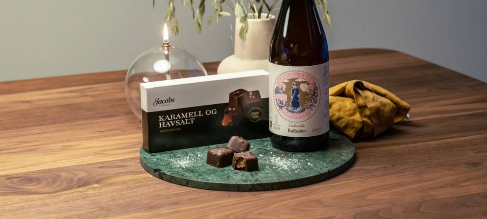 På et bord ligger en flaske med påskelogo, en liten eske med sjokolade, en lysestake med hvit lys og en gul duk, med en plante i bakgrunnen.