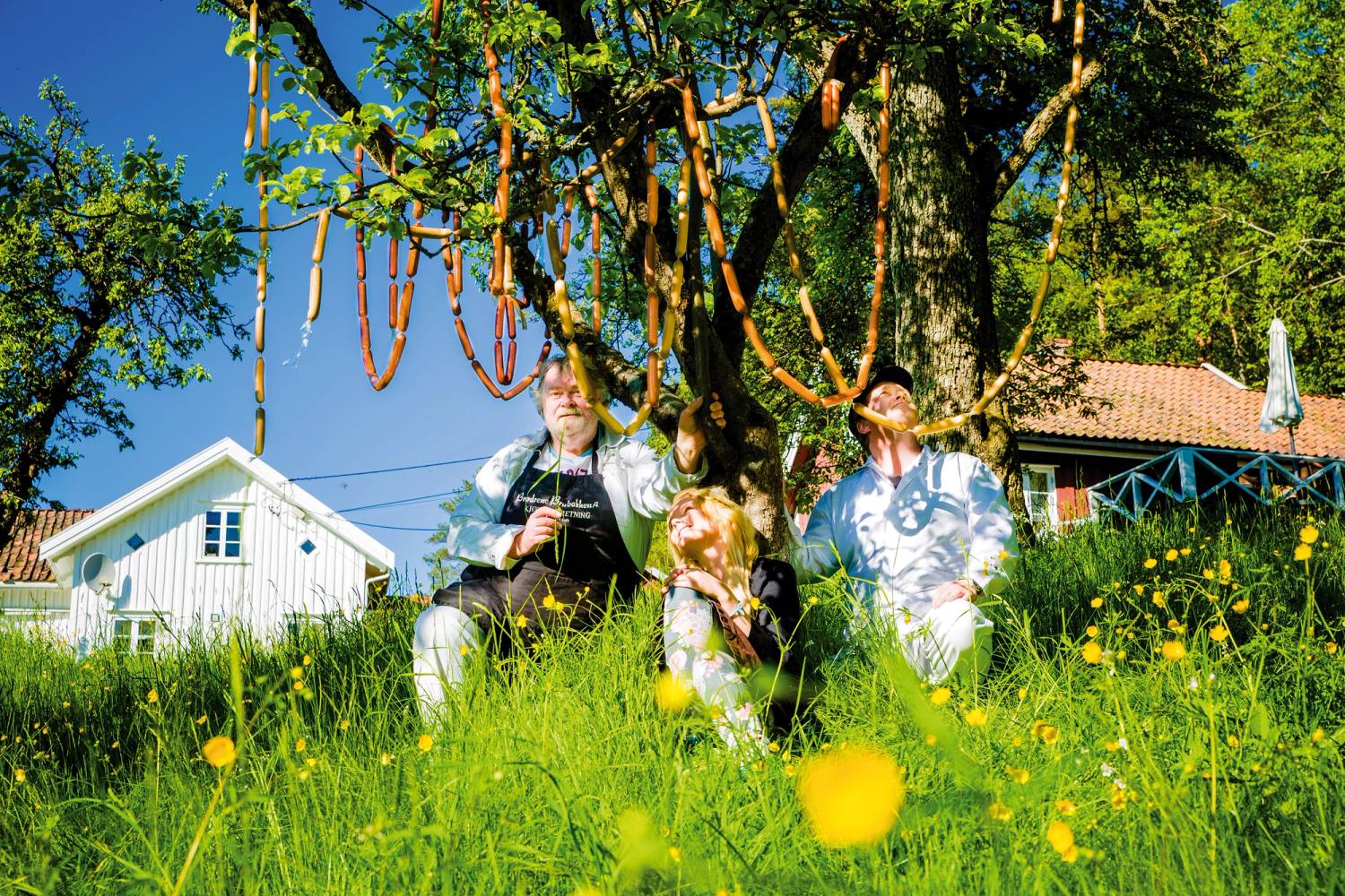 To personer sitter under et tre i en grønn hage med blomster, med en hvit hus i bakgrunnen og blå himmel over.
