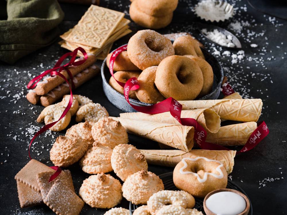 Julekaker og småkaker på en svart overflate med noe mel og julekrydder rundt, pyntet med røde bånd.