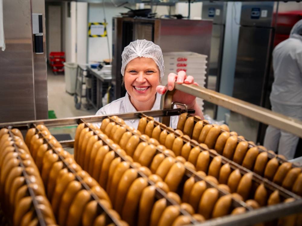 En person med hvitt hår som bruker vernebriller sjekker en metervare av brune kaker i en bakeri- eller produksjonsmiljø.