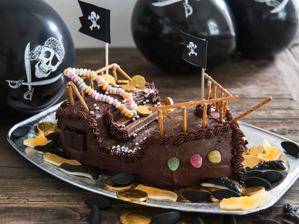 En sjørøverskute laget av sjokoladekake med små fargede godterier som dekorasjon, omgitt av svarte og hvite piratballonger med korsflagg.