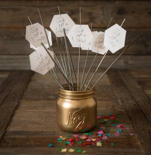 Små hvite kort med hilsener og lykkeønskninger festet til pinner i en gullfarget krukke, omgitt av fargerikt konfetti på et treverkebord.