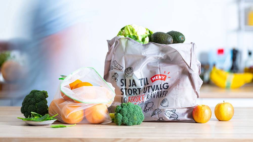 En gjenbrukspose fylt med grønnsaker og frukt på kjøkkenbenken, med en blå klesplagg i bakgrunnen.