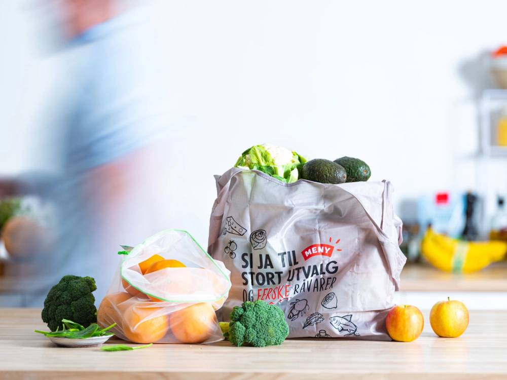 En gjenbrukspose fylt med grønnsaker og frukt på kjøkkenbenken, med en blå klesplagg i bakgrunnen.