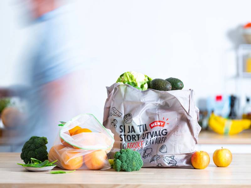 En gjenbrukspose fylt med grønnsaker og frukt på kjøkkenbenken, med en blå klesplagg i bakgrunnen.