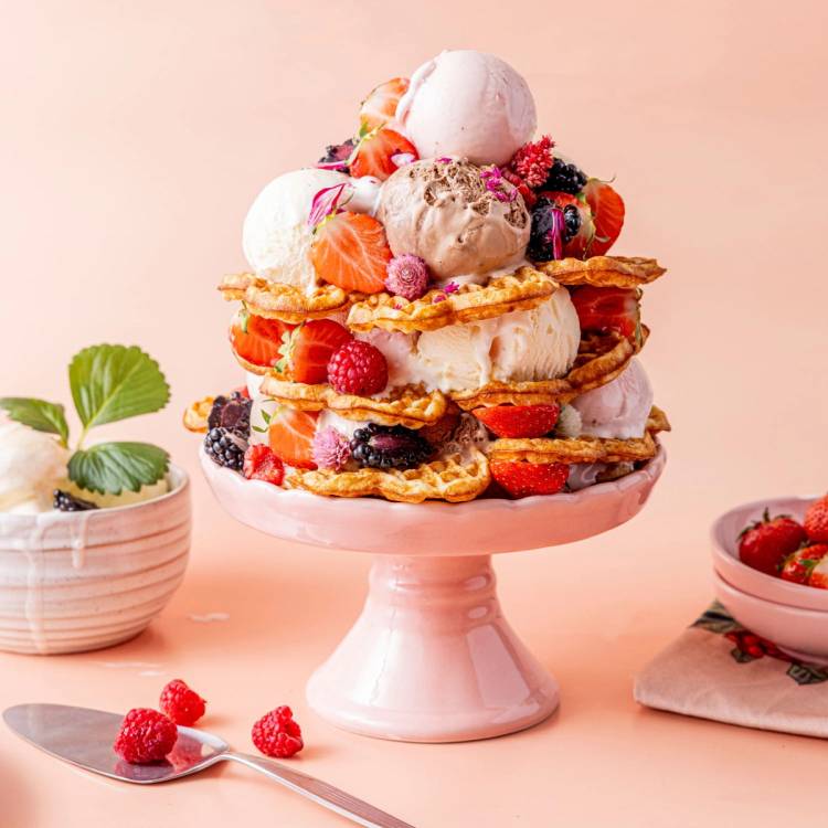 Vaffeltårn med iskrem og bær på en rosa kakefat, omgitt av små boller med bær og en skje med noen bær, på et lyst bord.