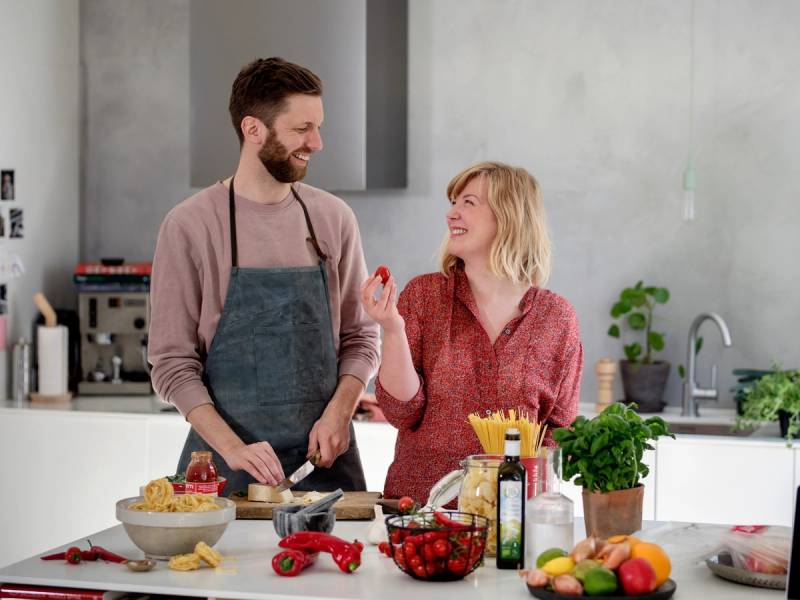 To personer i kjøkkenet som lager mat sammen, med ulike ingredienser og kjøkkenutstyr på benken.