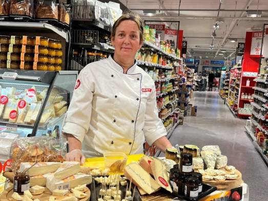 En person i hvit kokkeuniform står i en matbutikk med ost og andre meieriprodukter på bordet foran seg.