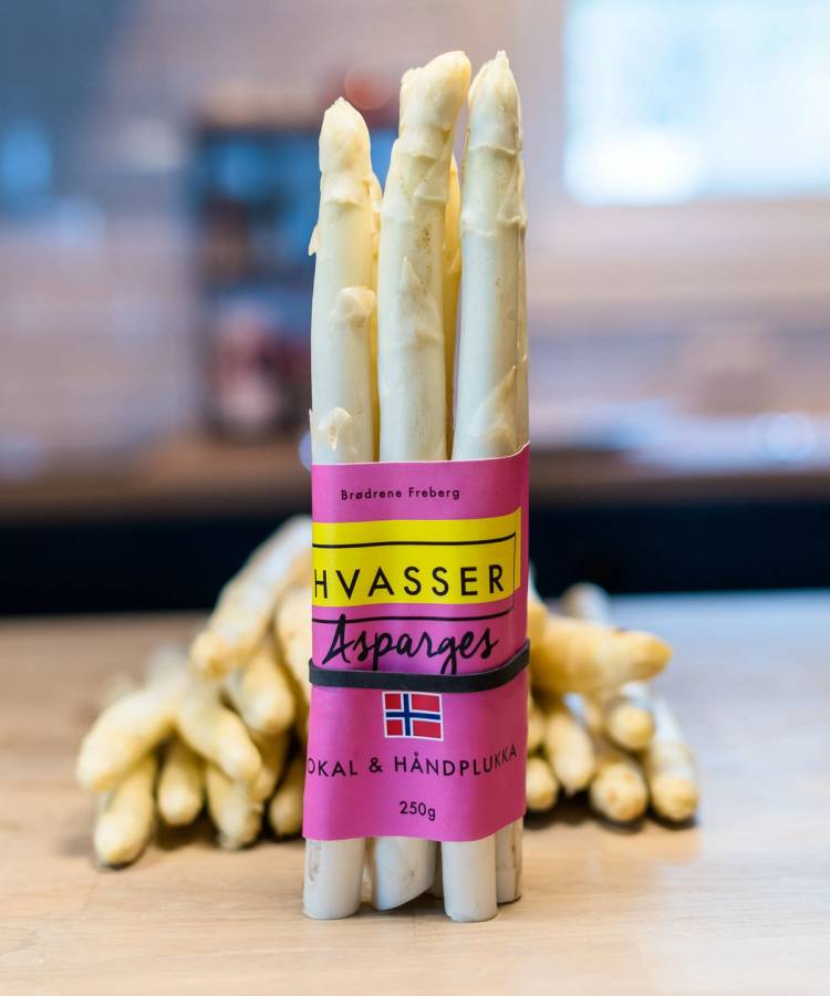 Pakke med hvit asparges med rosa og gul etikett som sier 'Hvasser Asparges' og norsk flagg, plassert på et bord med flere asparges i bakgrunnen.