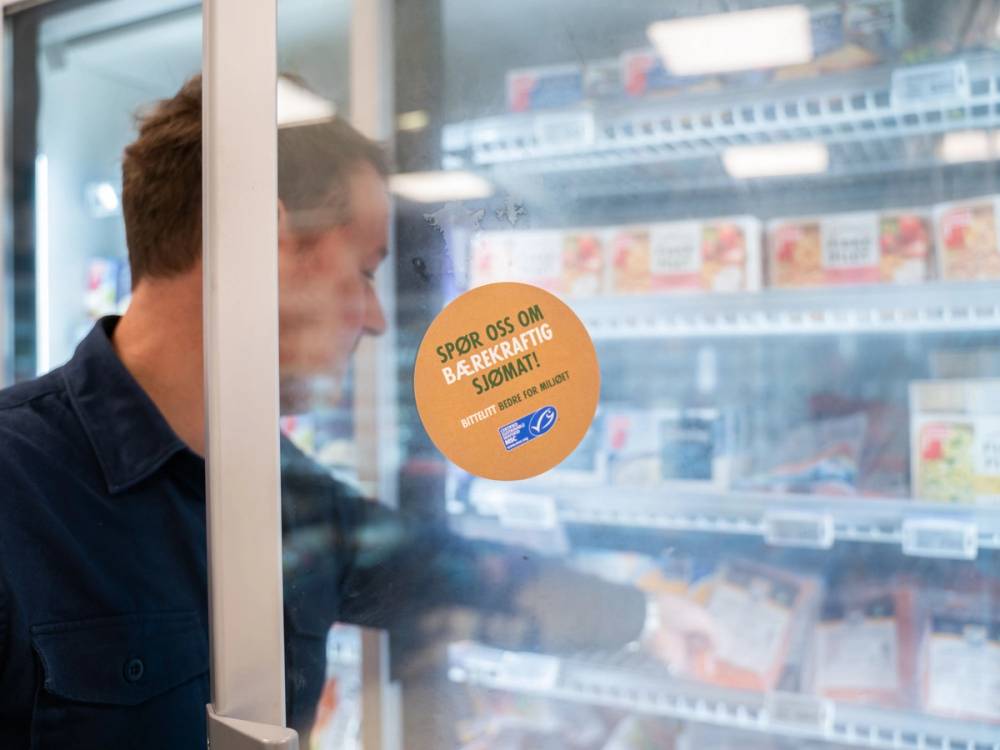 En person ser på kjøttprodukter i en kjøledisk på en dagligvarebutikk, med en reklameskilt på glasset.