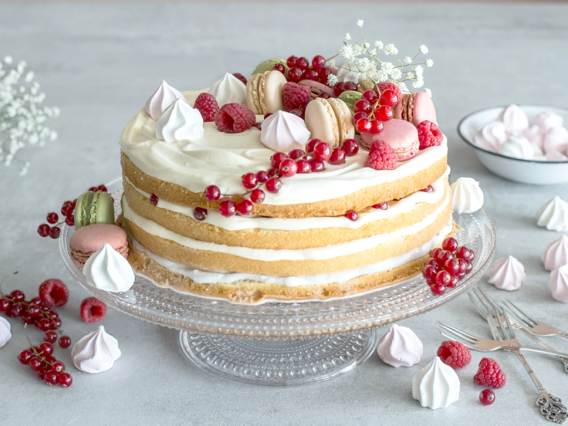 En sommerbløtkake med krem, bær og makroner på en glasskakefat, omgitt av små kaker og bær på et lyst bord.
