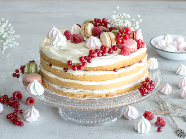 En sommerbløtkake med krem, bær og makroner på en glasskakefat, omgitt av små kaker og bær på et lyst bord.
