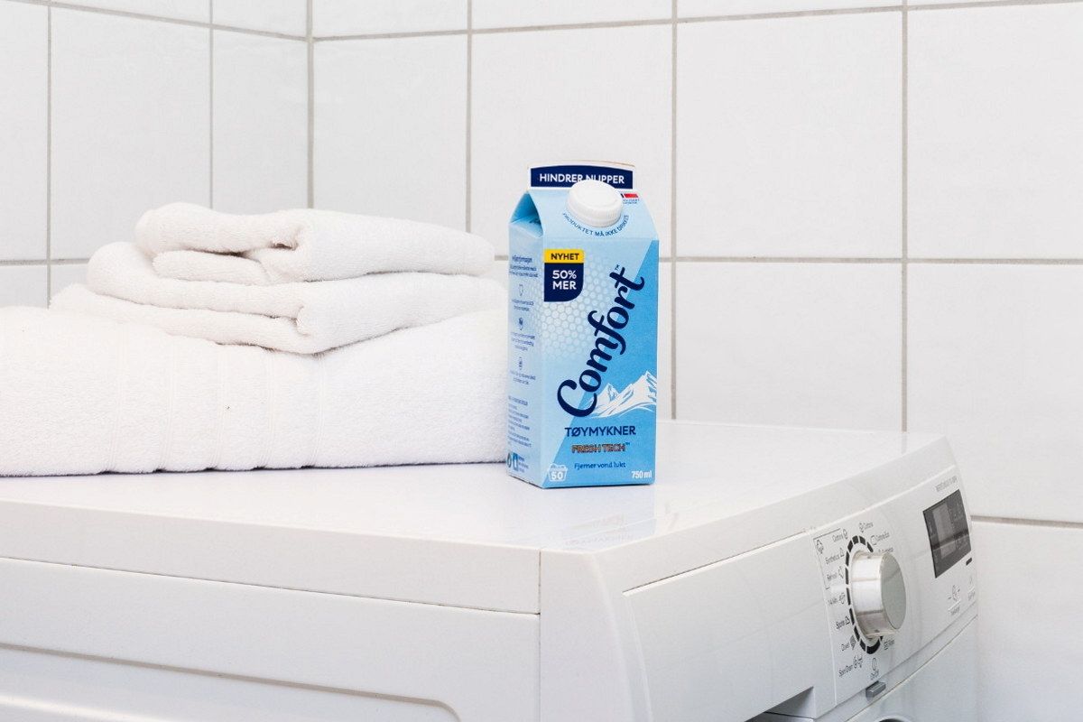 En vaskemaskin med en kartong Comfort-blekemiddel og noen håndklær på toppen i et bad eller vaskerom.