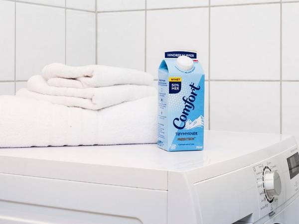 En vaskemaskin med en kartong Comfort-blekemiddel og noen håndklær på toppen i et bad eller vaskerom.