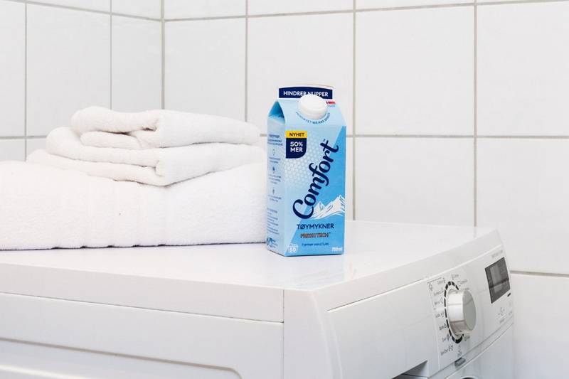 En vaskemaskin med en kartong Comfort-blekemiddel og noen håndklær på toppen i et bad eller vaskerom.