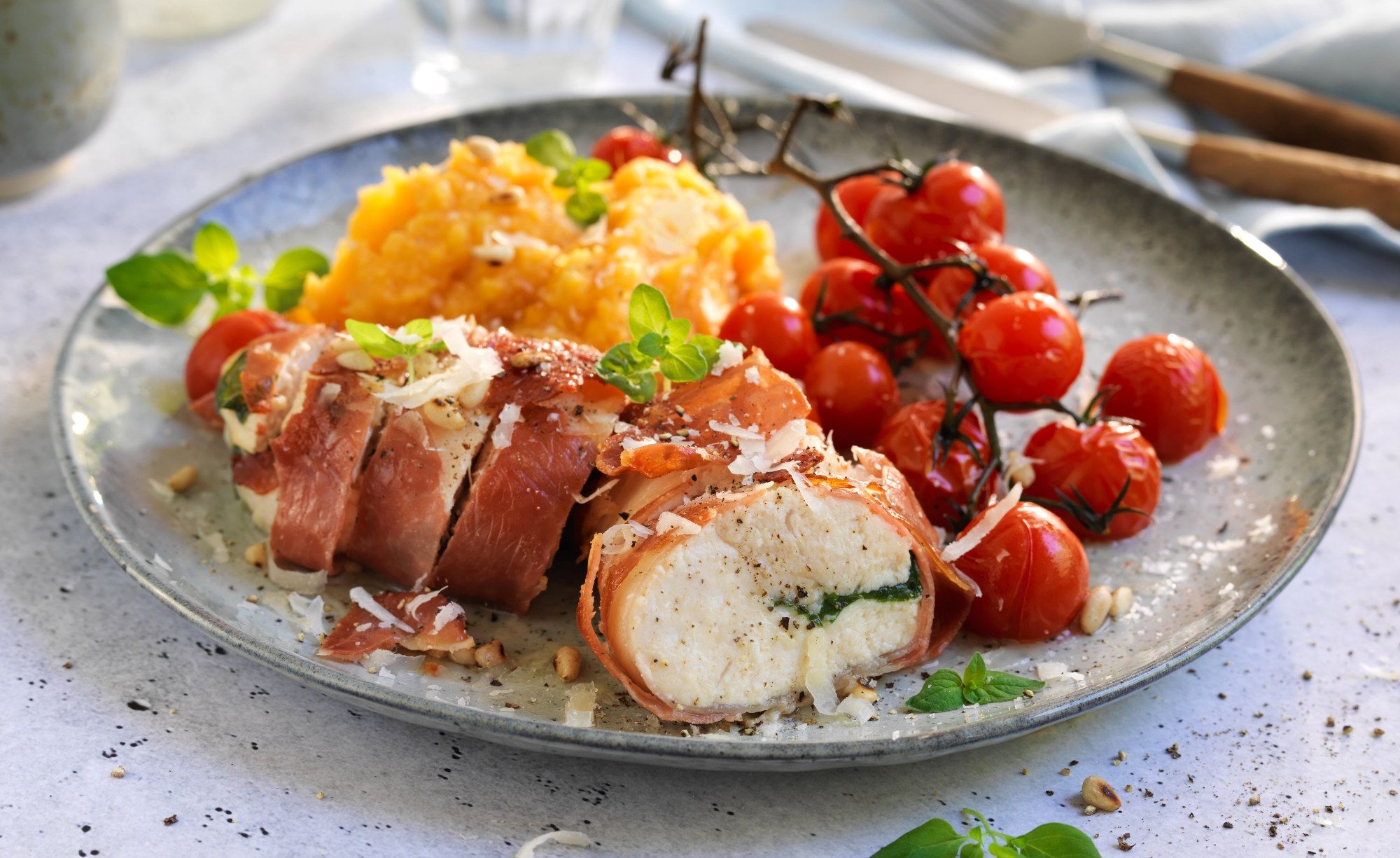 Kyllingbryst fylt med manchago ost og surret i spekeskinke, servert med potetmos og cherrytomater på en tallerken.