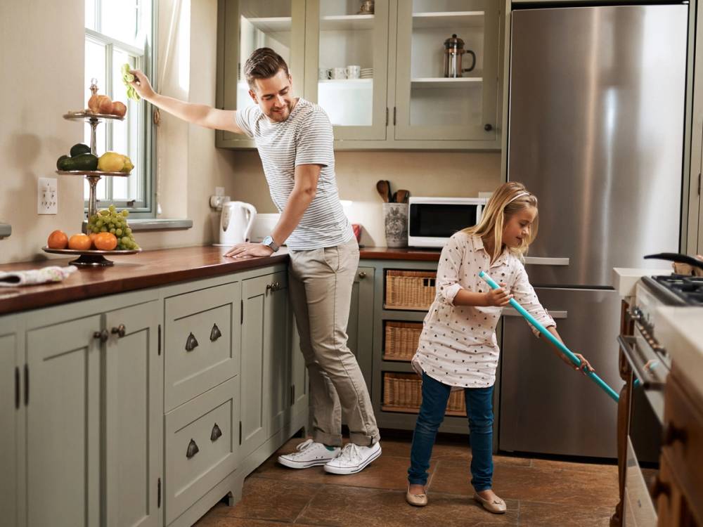 En voksen og et barn gjør rent på kjøkkenet med mopp og klut, mens de står ved kjøkkenbenken og kjøleskapet.