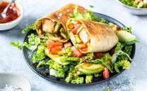 Tofuwrap med grønnsaker på en svart tallerken, omgitt av små skåler med saus og salat.