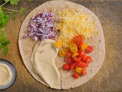 En tortilla wrap med oppskåret cherrytomater, revet ost, hakket rødløk og en klatt med saus eller hummus på en treplate.