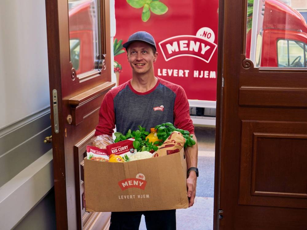 Person som leverer en stor eske med mat ut av en butikkdør, med meny-logo i bakgrunnen.