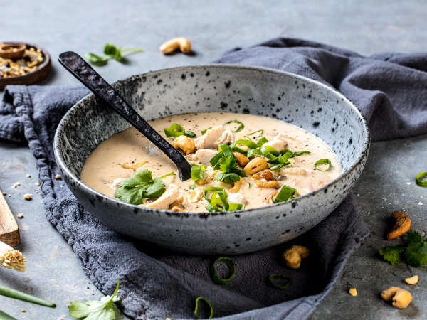 En bol med thai-kyllingsuppe pyntet med grønne løk og peanøtter, plassert på en mørk duk med noen urter og nøtter rundt.