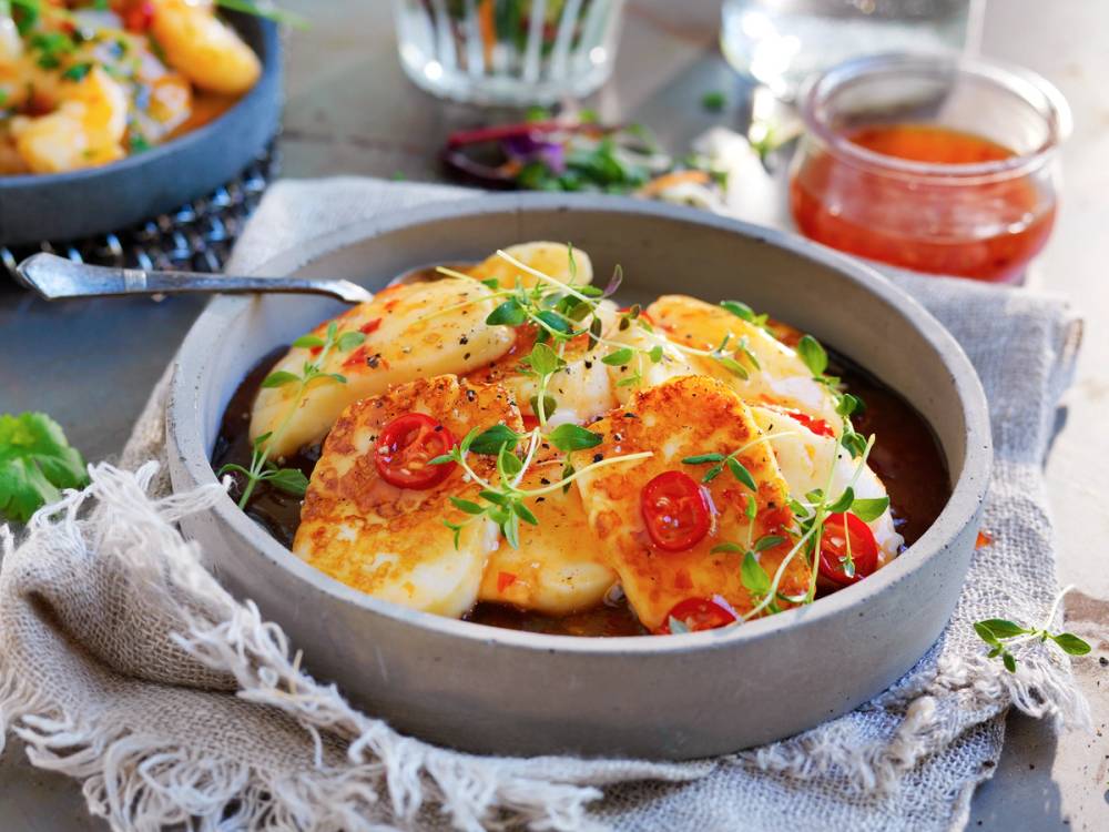 Stekt halloumi servert med cherrytomater og urter i en grå serveringsskål, med en liten skål med saus i bakgrunnen.