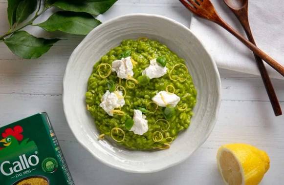 Risotto med grønne erter og små biter av hvit ost i en hvit bolle på et lyst bord med skjeer og sitron i nærheten.