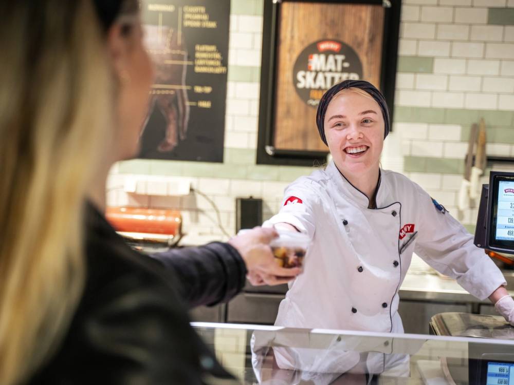 En kunde mottar mat fra en ansatt bak disken på en matbutikk eller restaurant, med meny og prislister i bakgrunnen.