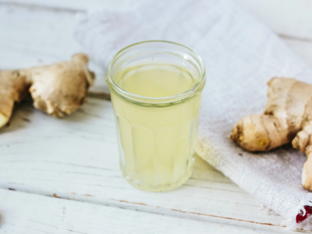 Et glass med ingefærshot på et hvitt bord med ingefærrot i bakgrunnen