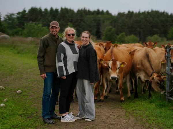 Tre personer står sammen i en åker med kyr i bakgrunnen, og en av personene har en hånd på en ku.