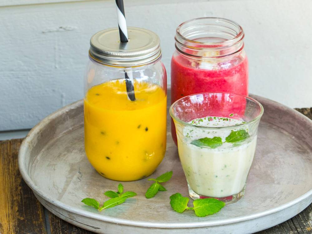 Tre friske smoothies i glass med ulike farger på en rund tallerken, pyntet med mynteblader.